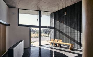 Interior view of Crematorio Igualada with light-bulb style ceiling lights hanging from the ceiling. An oak bench against a grey wall with vertical lines by a floor to ceiling window. Grey flooring and concrete coloured ceilings