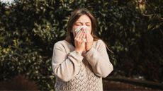 Woman sneezing into tissue standing next to tree, representing a hay fever cough
