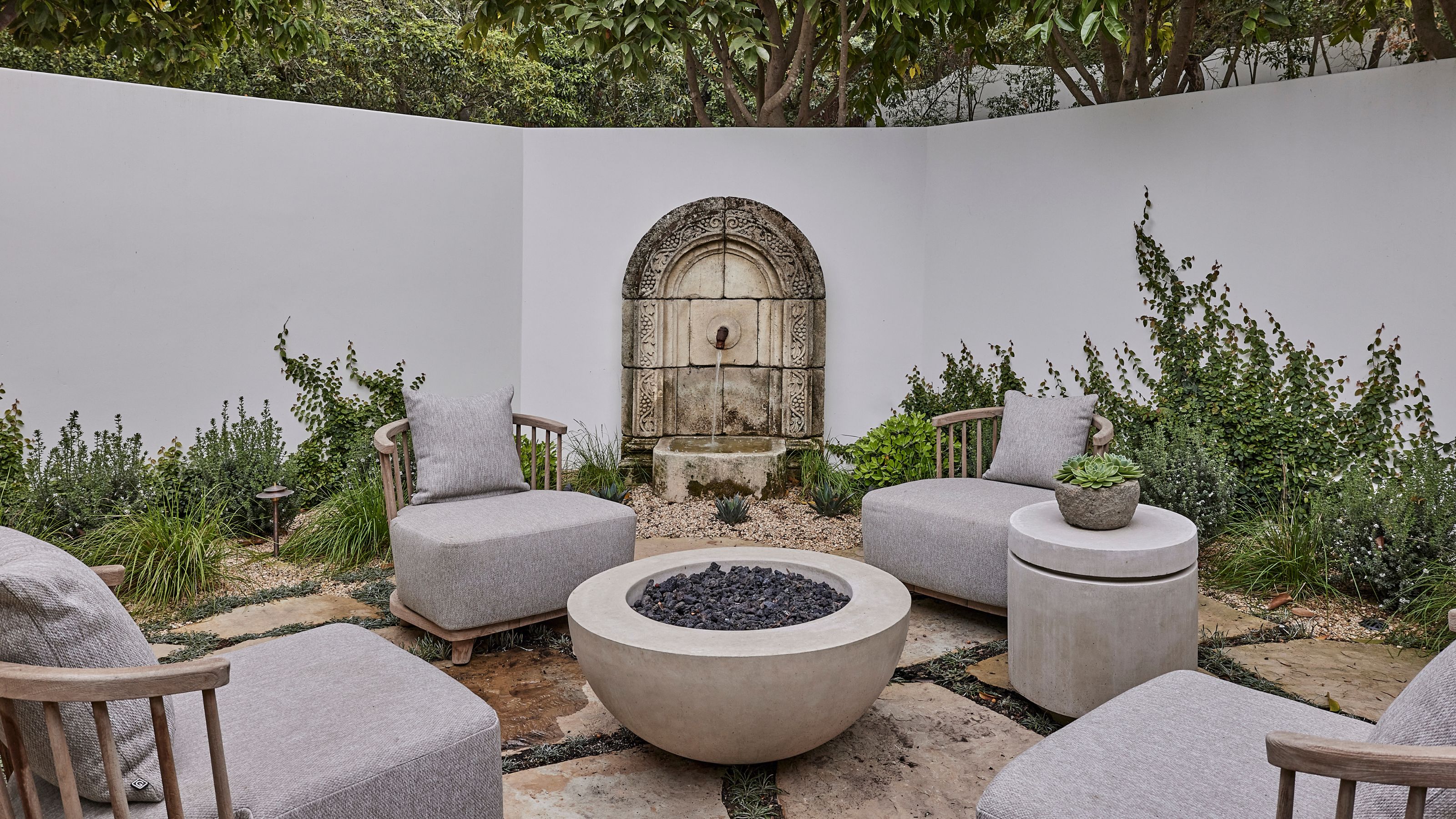 a walled spanish-style courtyard garden seating area with a stone water feature, stone flooring and firepit at the center