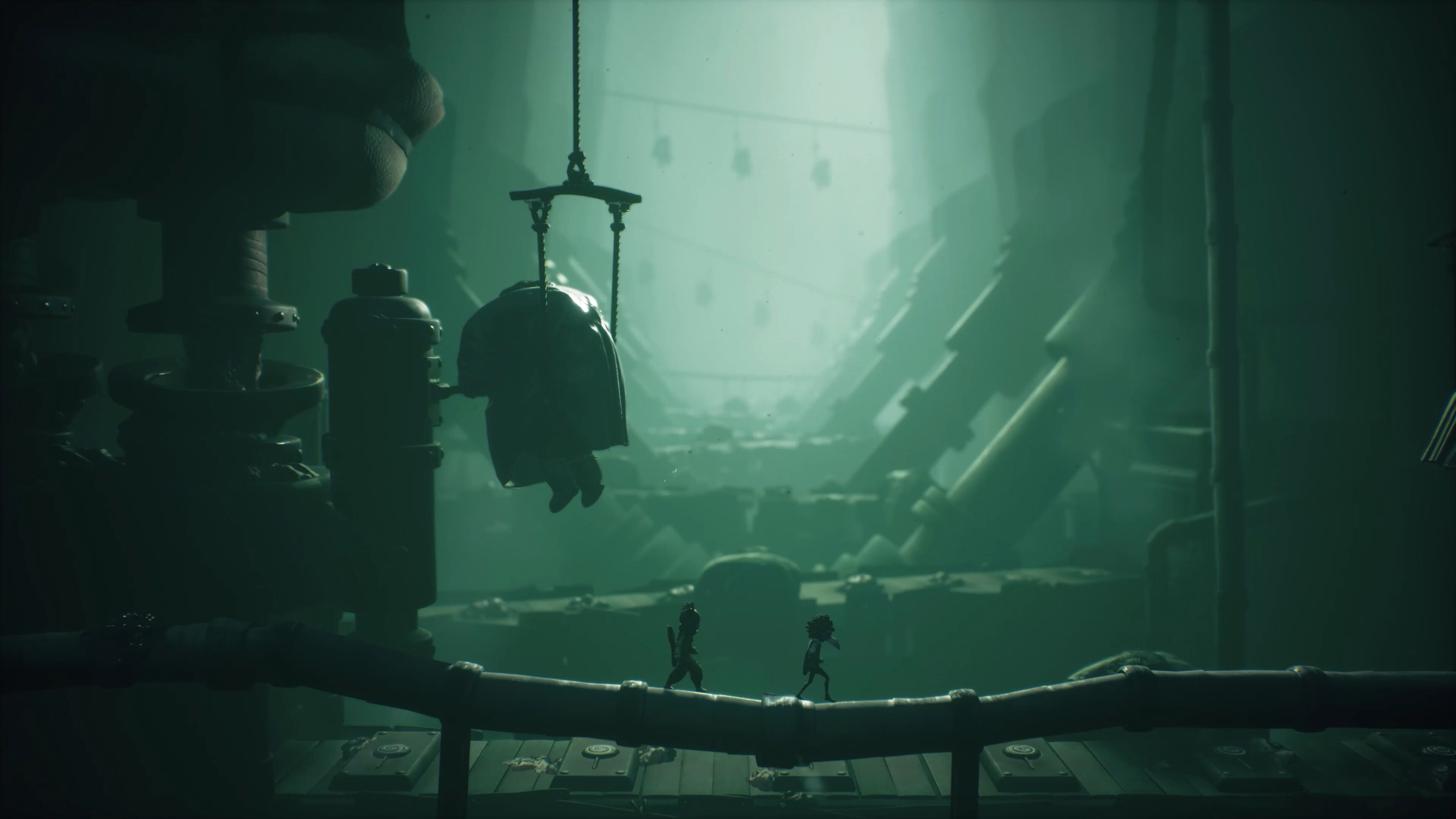 Alone and Low walk across a pipe through a green-tinged room in Little Nightmares 3 with a large figure in a sling in the background working machinery