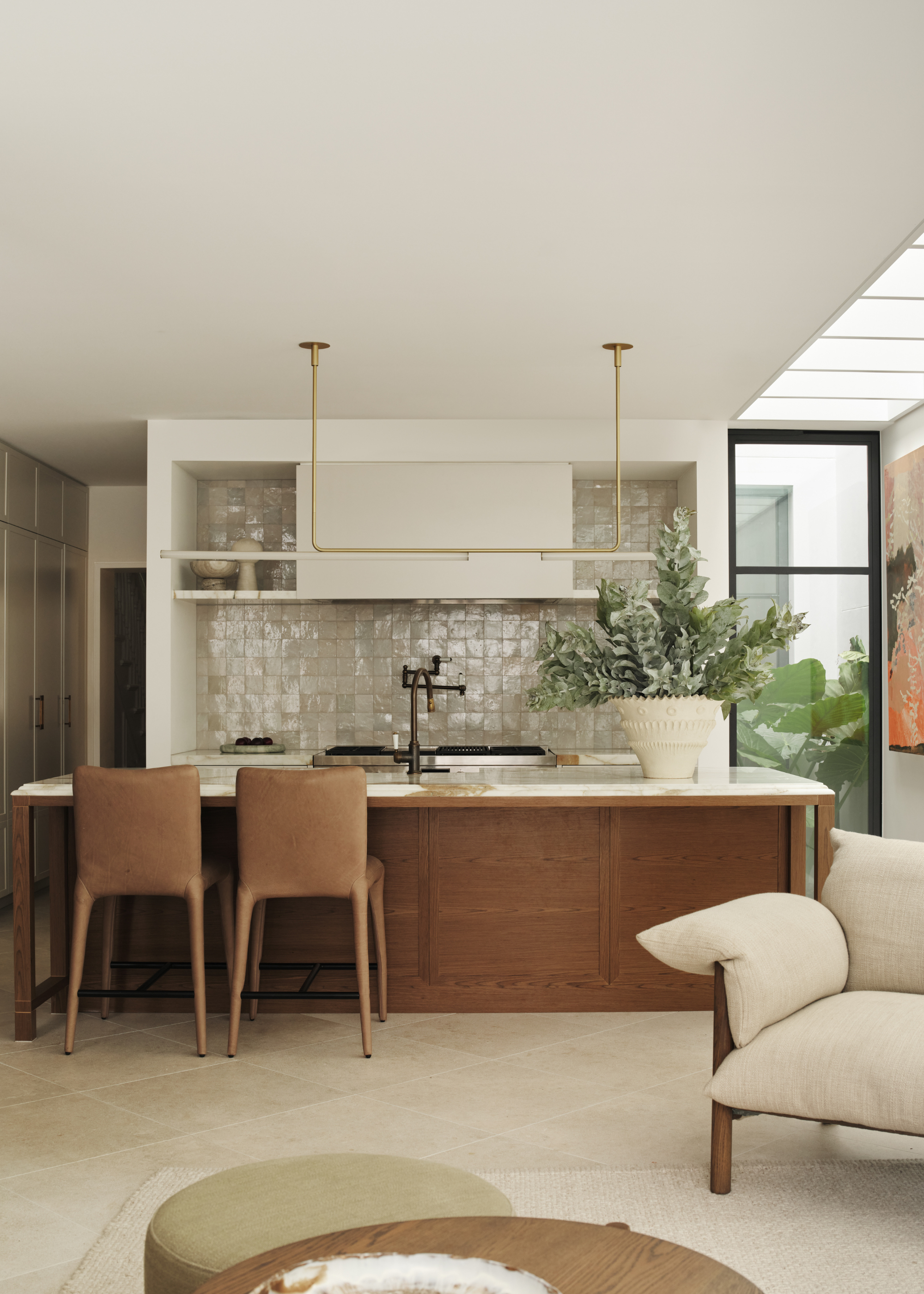 A bright kitchen with a stone and wood island, a large vase of stems, a set of tan chairs, a zellige tile backsplash and a gold rung