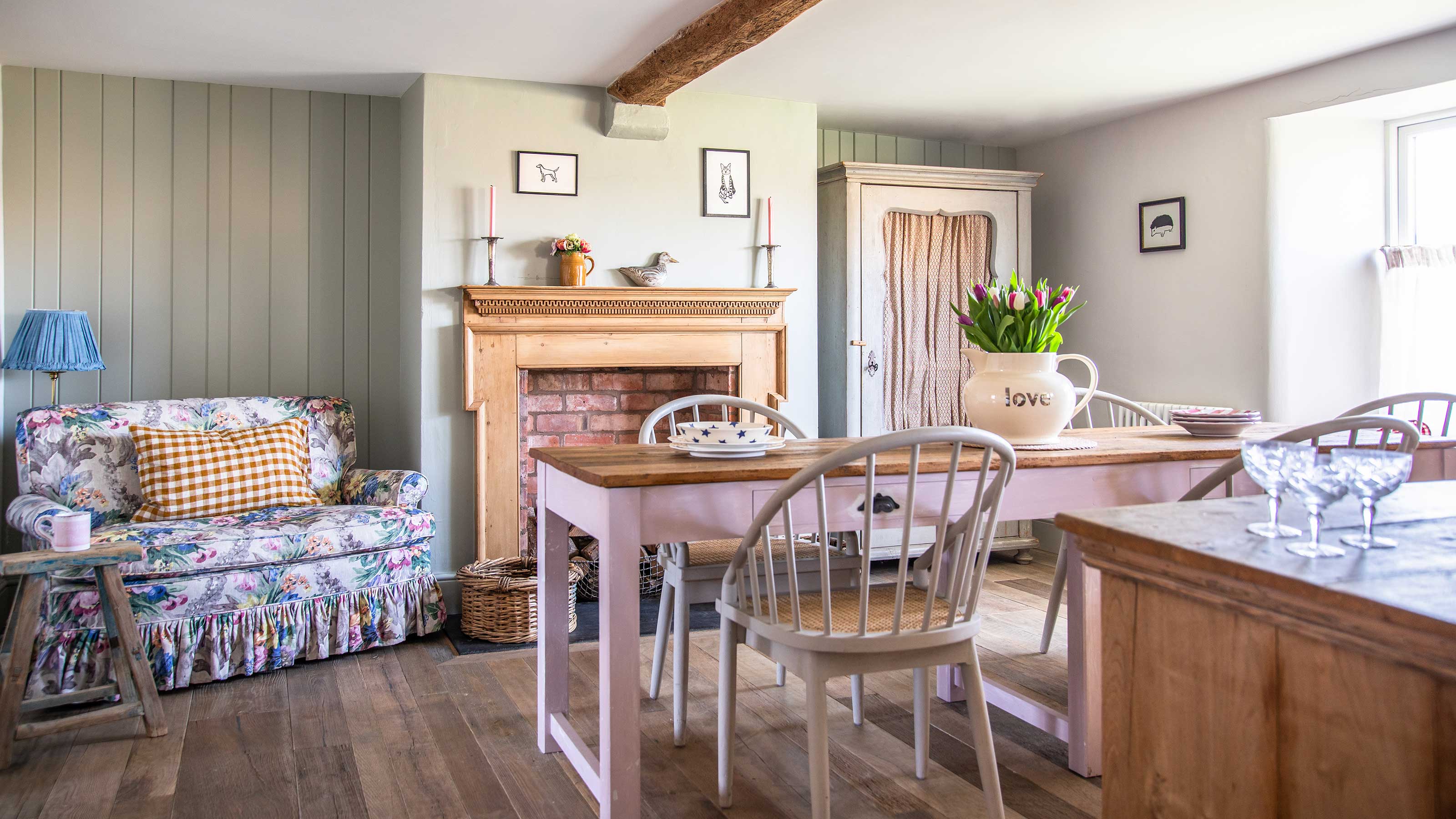 country dining room with pink painted farmhouse table, wooden mantelpiece, floral sofa