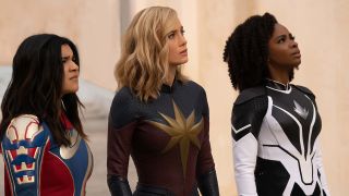 Still from The Marvels (2023) movie. All The Marvels all standing together looking upwards in the same direction. From left to right: Ms. Marvel (teen with long dark hair wearing a red, white and blue supersuit), Captain Marvel (long blonde hair wearing a blue and red supersuit with a giant gold star-like emblem on the front) and Monica Rambeau (dark tightly curled hair down to her shoulders and wearing a black and white supersuit with a smaller silver teardrop-shaped emblem on her chest).