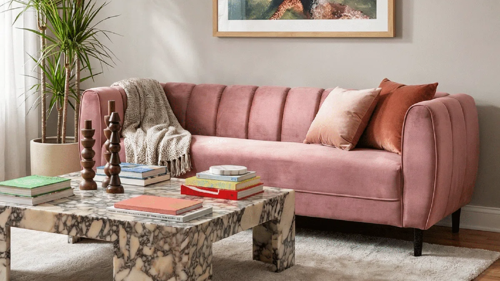 a living room with a pink couch, a marble coffee table, pillows, books, candle holders, and a large plant in the left corner
