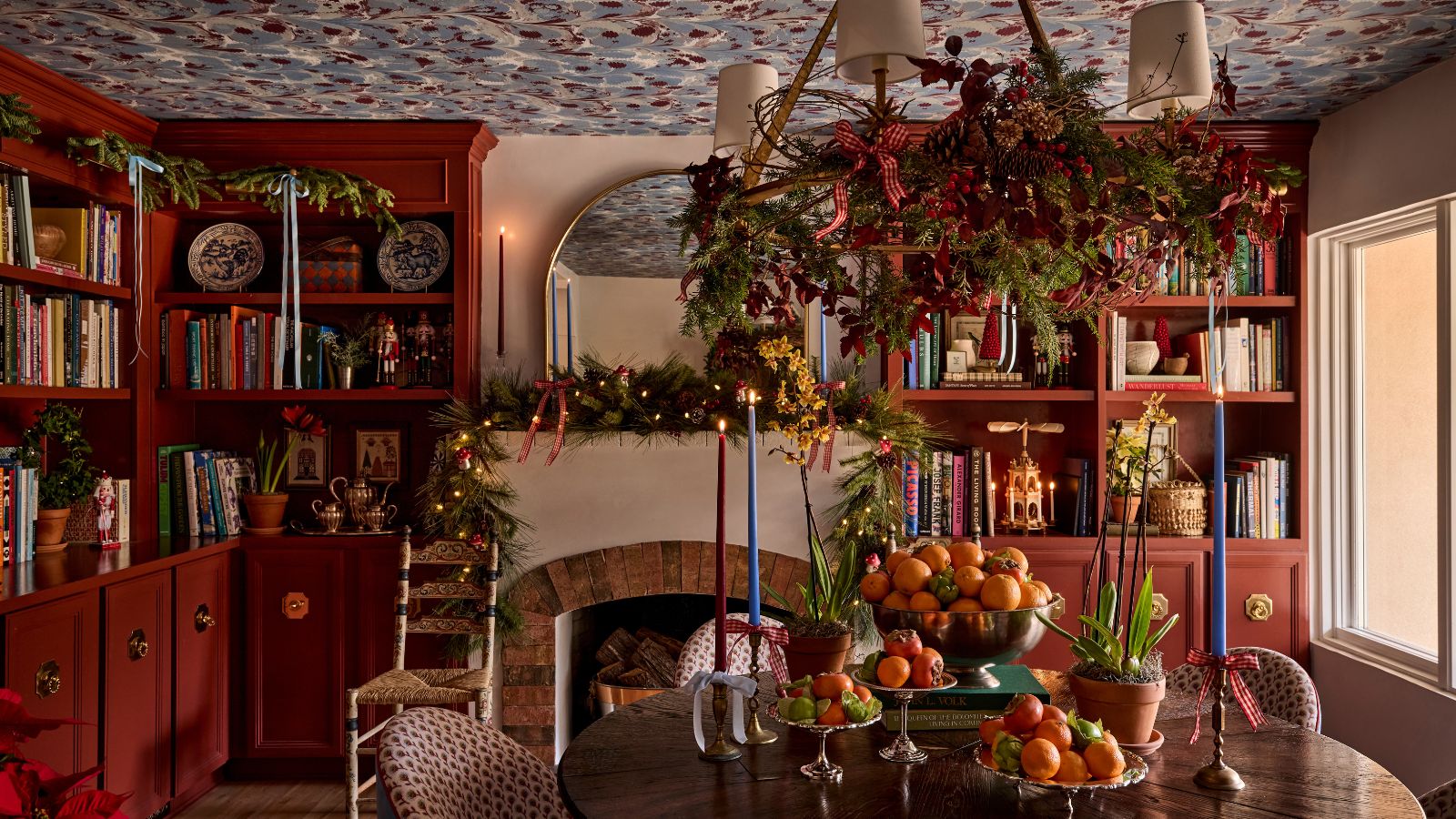 Dining room with red built-in bookcases, a marble blue and red ceiling, a fireplace with a festive garland on the mantel, and a dining table decorated with bowls of clementines and taper candles.