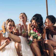 A bride and three bridesmaids sitting together on the beach laughing with flowers in their hand