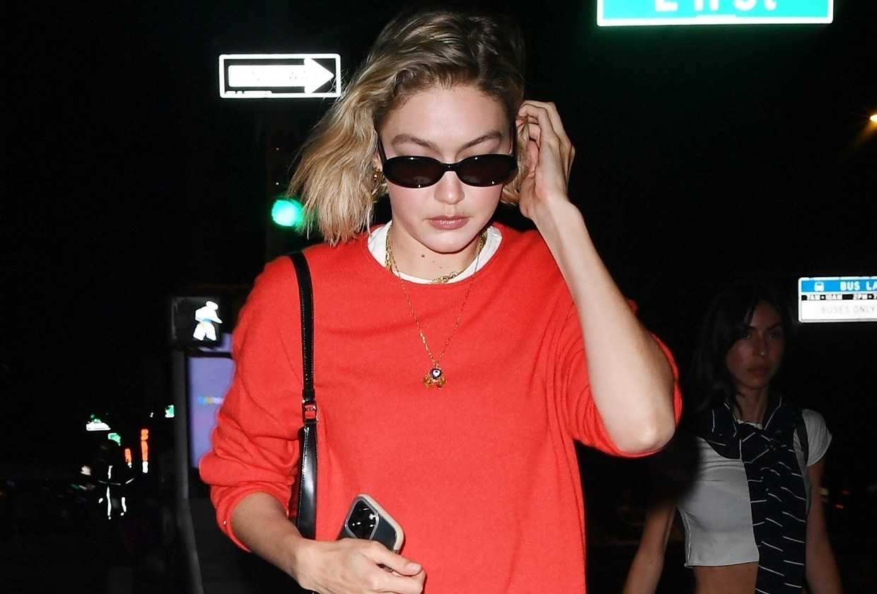Gigi Hadid walking in New York City wearing a red T-shirt, sunglasses, sneakers, and a white eyelet skirt.