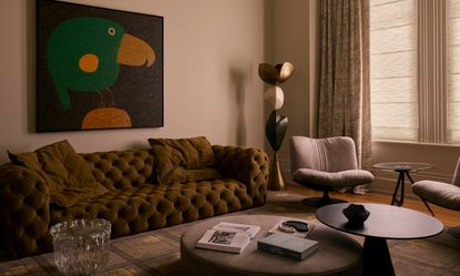 Living room with light beige walls, khaki sofa, toucan artwork, taupe velvet ottoman and ceramic totem-style sculpture