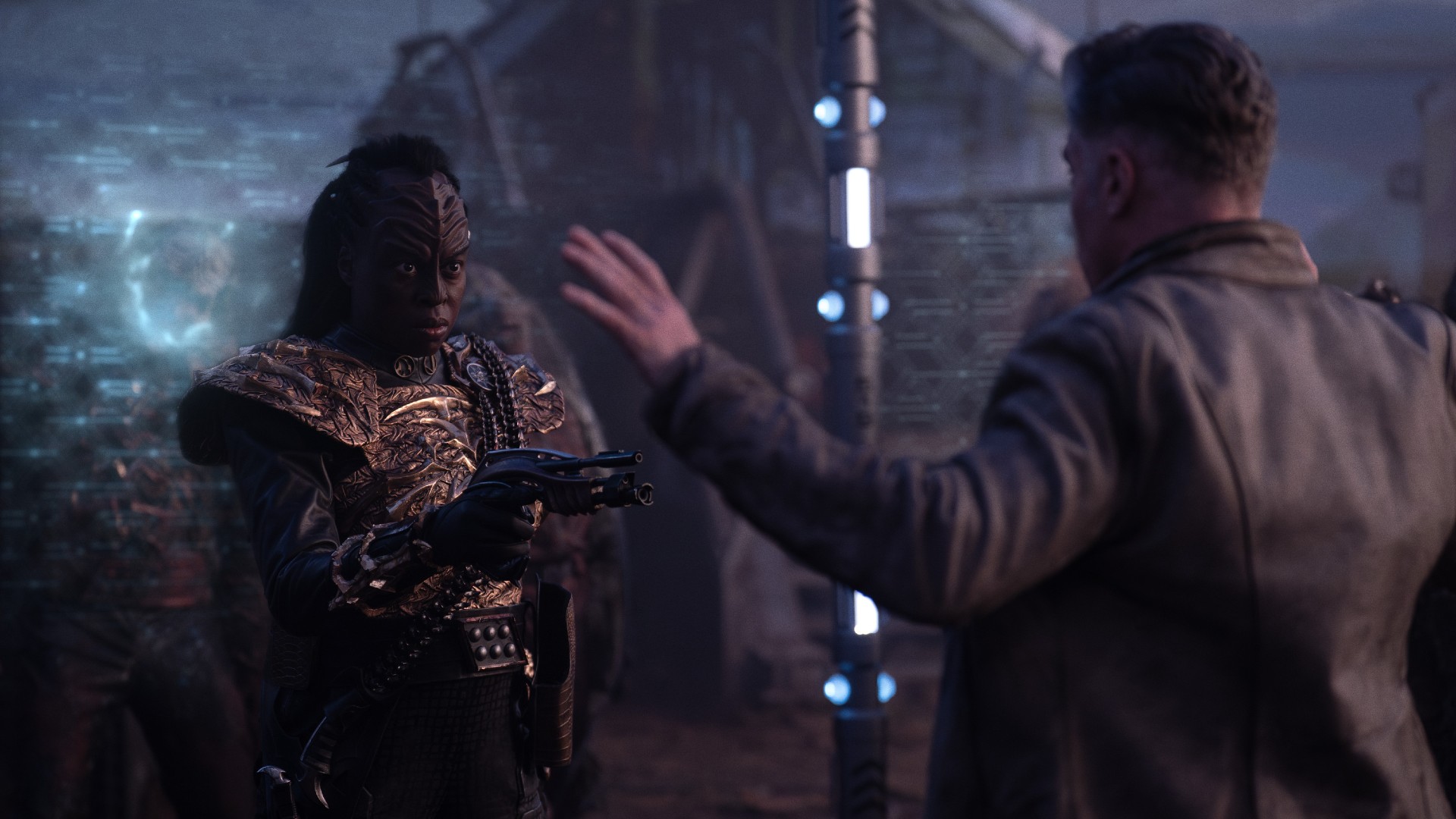 An angry female Klingon is pointing a gun at a man (he is holding his hands up in surrender). Behind them are zombies are held off by a forcefield. L to R Christine Horn as Bytha and Anson Mount as Capt. Pike in season 3, Episode 3 of Strange New Worlds streaming on Paramount+.