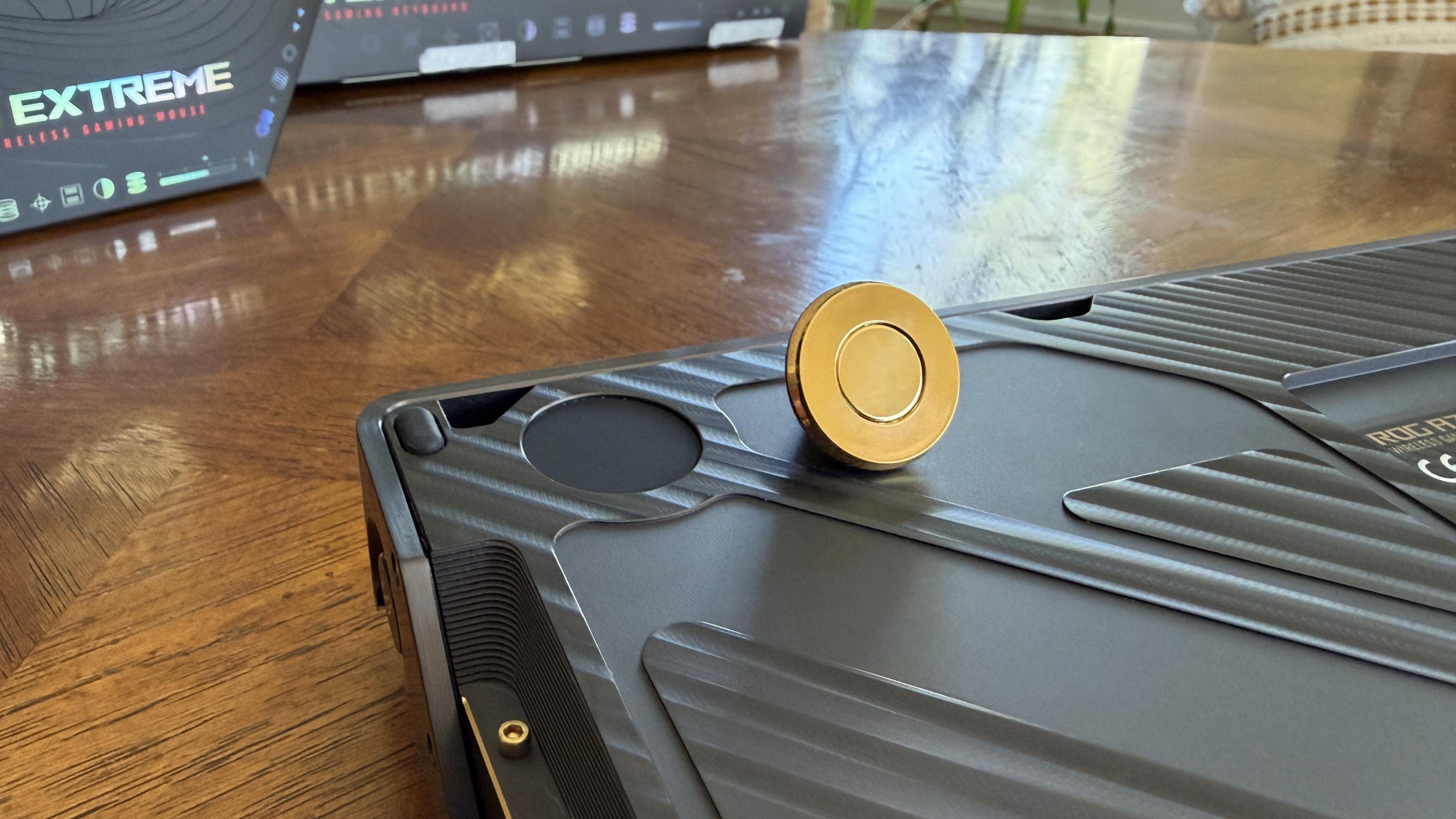 Close-up of a dark gray ASUS ROG Azoth Extreme Edition 20 with a gold circular object on a wooden surface, and a box labeled "EXTREME" in the background.