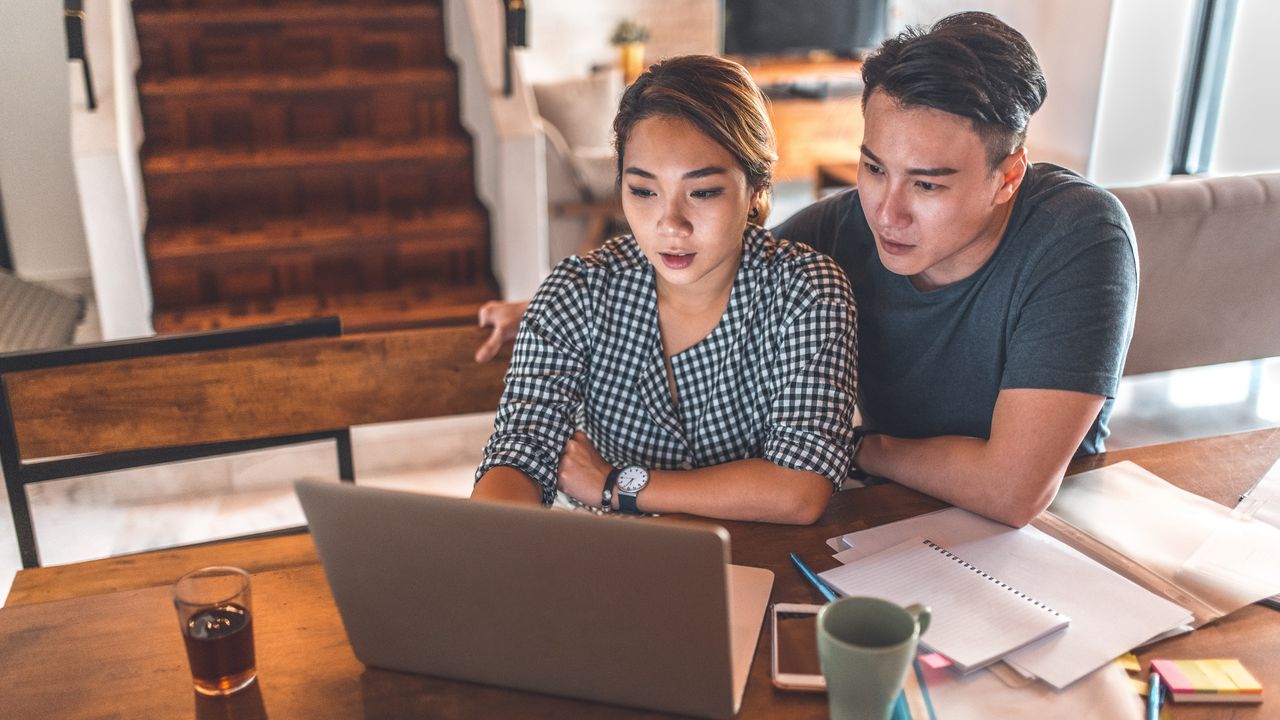 A young couple work together on a laptop with paperwork strewn around them.