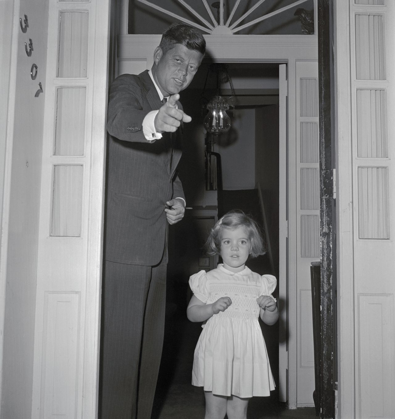 President-elect John F. Kennedy seems to be pointing to newsmen gathered outside his Georgetown home, as he poses with his daughter, Caroline, soon to be three-years-old