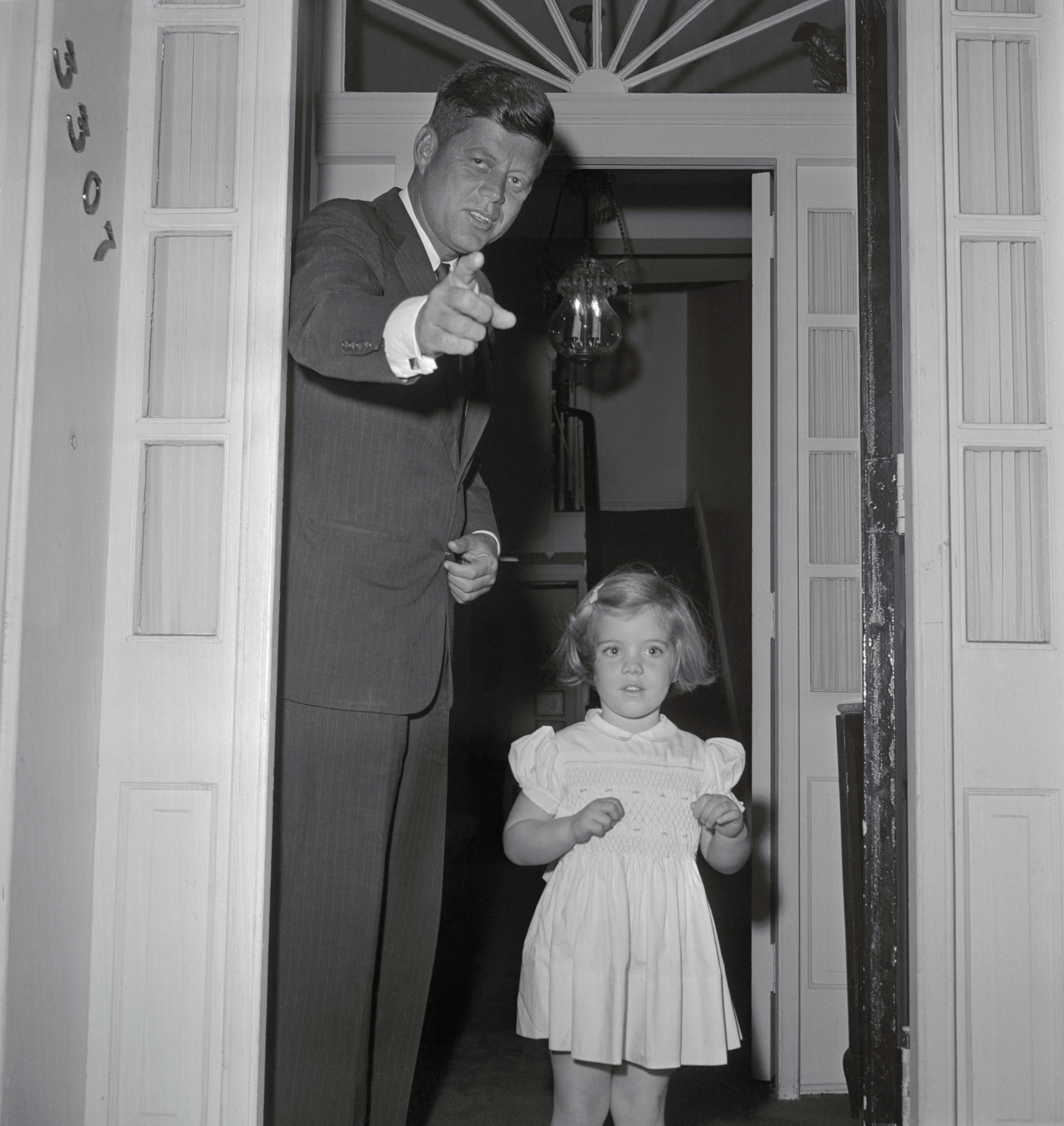 President-elect John F. Kennedy seems to be pointing to newsmen gathered outside his Georgetown home, as he poses with his daughter, Caroline, soon to be three-years-old