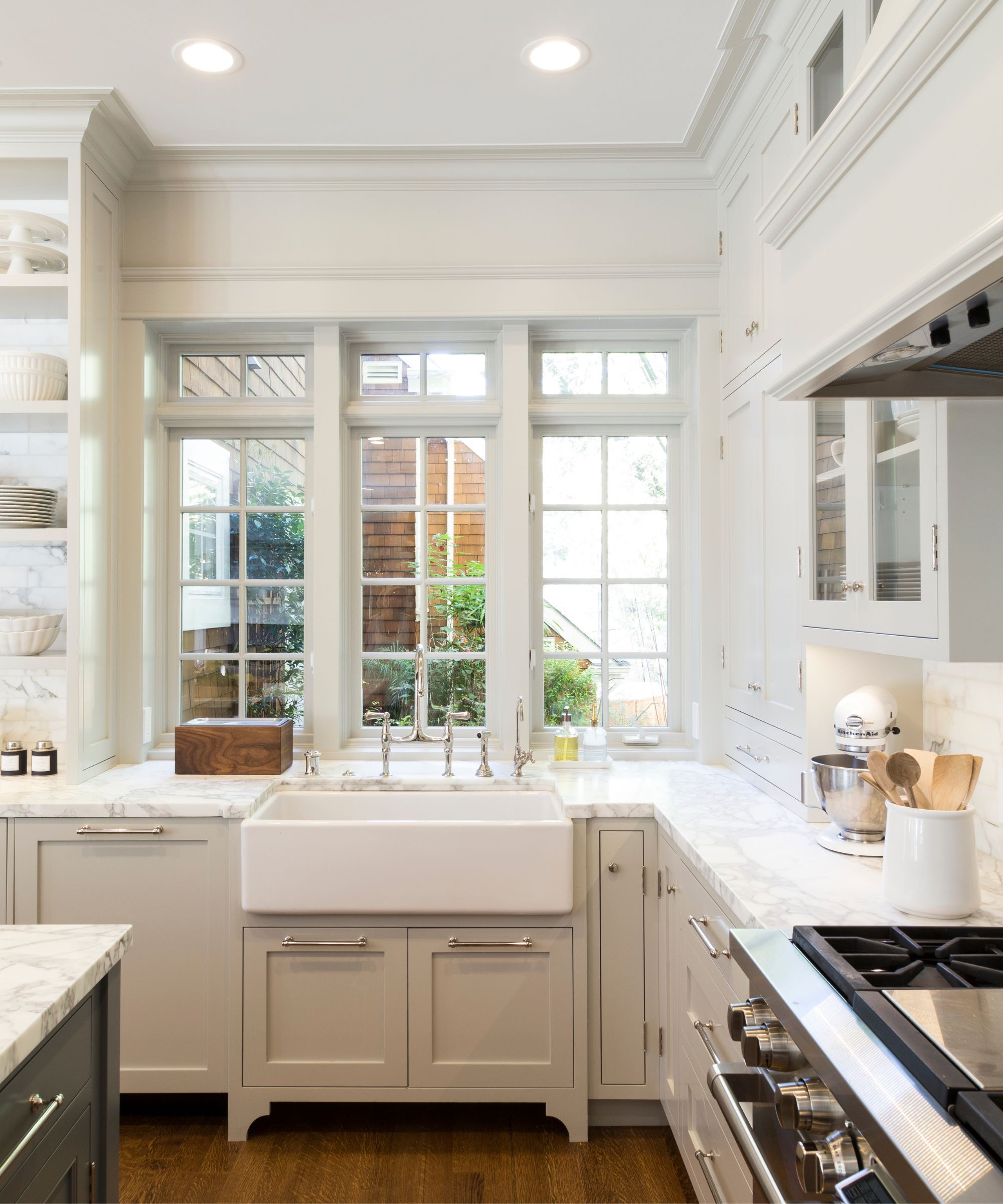 Bright kitchen with Gray Owl by Benjamin Moore painted cabinetry with glass fronts, white marble countertops, a silver range oven, a white sink, and a large window overlooking a garden.