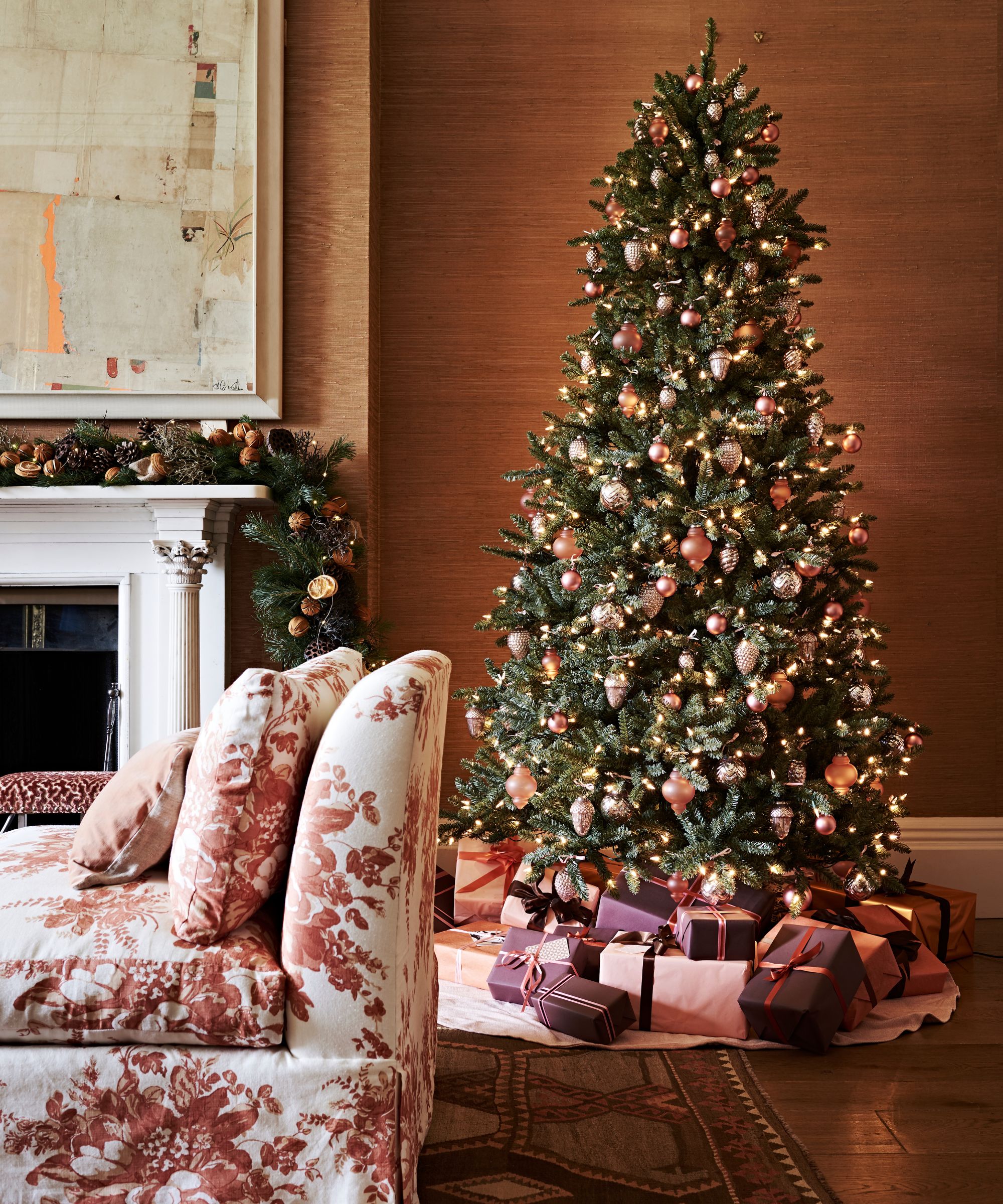 A festive living room with a large decorated tree with gifts beneath, and dark orange walls. To the left of the tree is a white mantelpiece with festive foliage and a large artwork above, and in the foreground is a white and red floral couch.