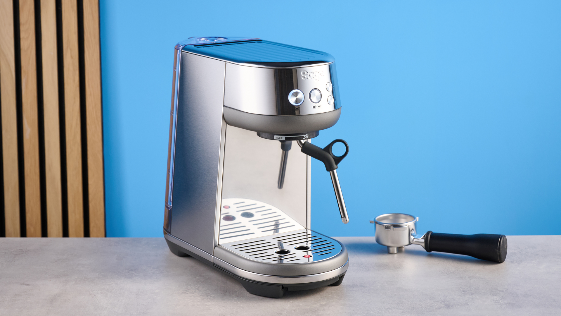 the breville bambino / sage bambino in silver photographed against a blue background with a wooden feature wall, showing the steam wand, group head, water tank, button controls, and espresso