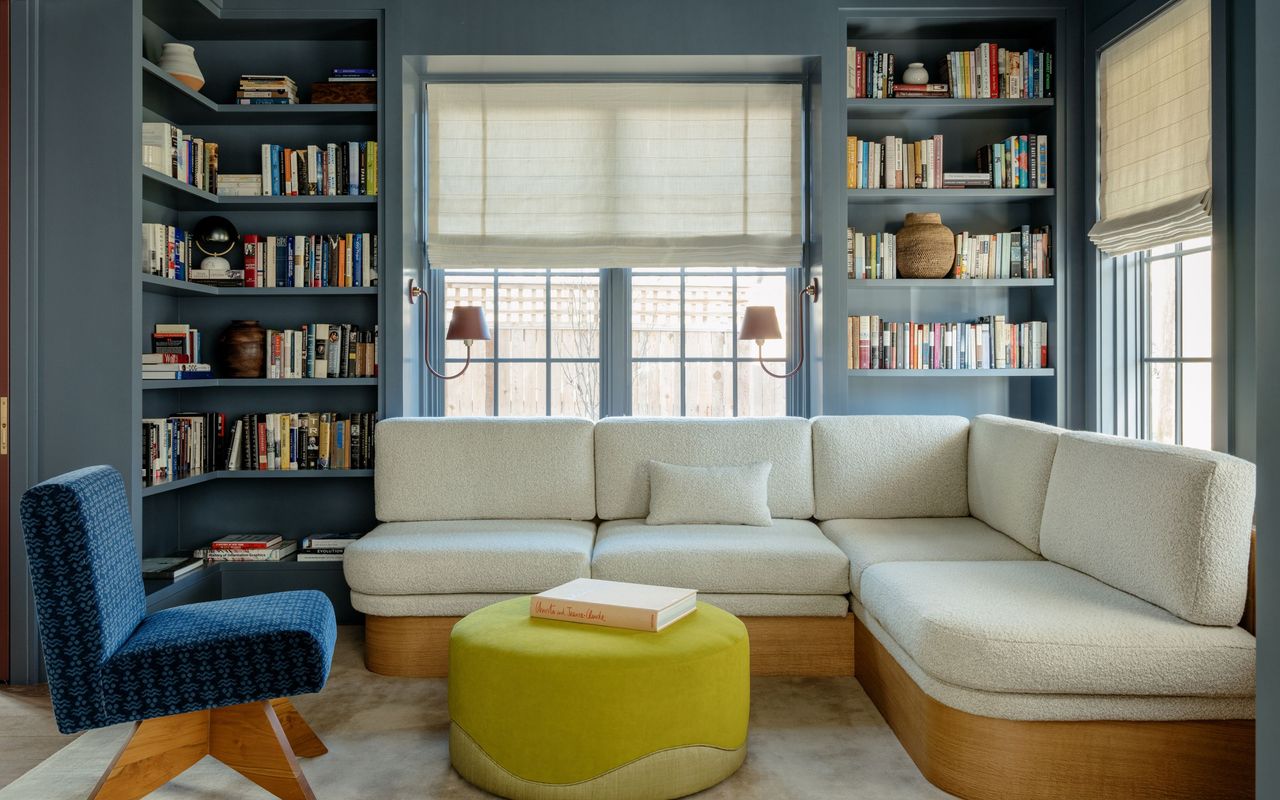 home library snug with blue walls and shelvesm light colored sofa, blue accent chair, and yellow pouffe