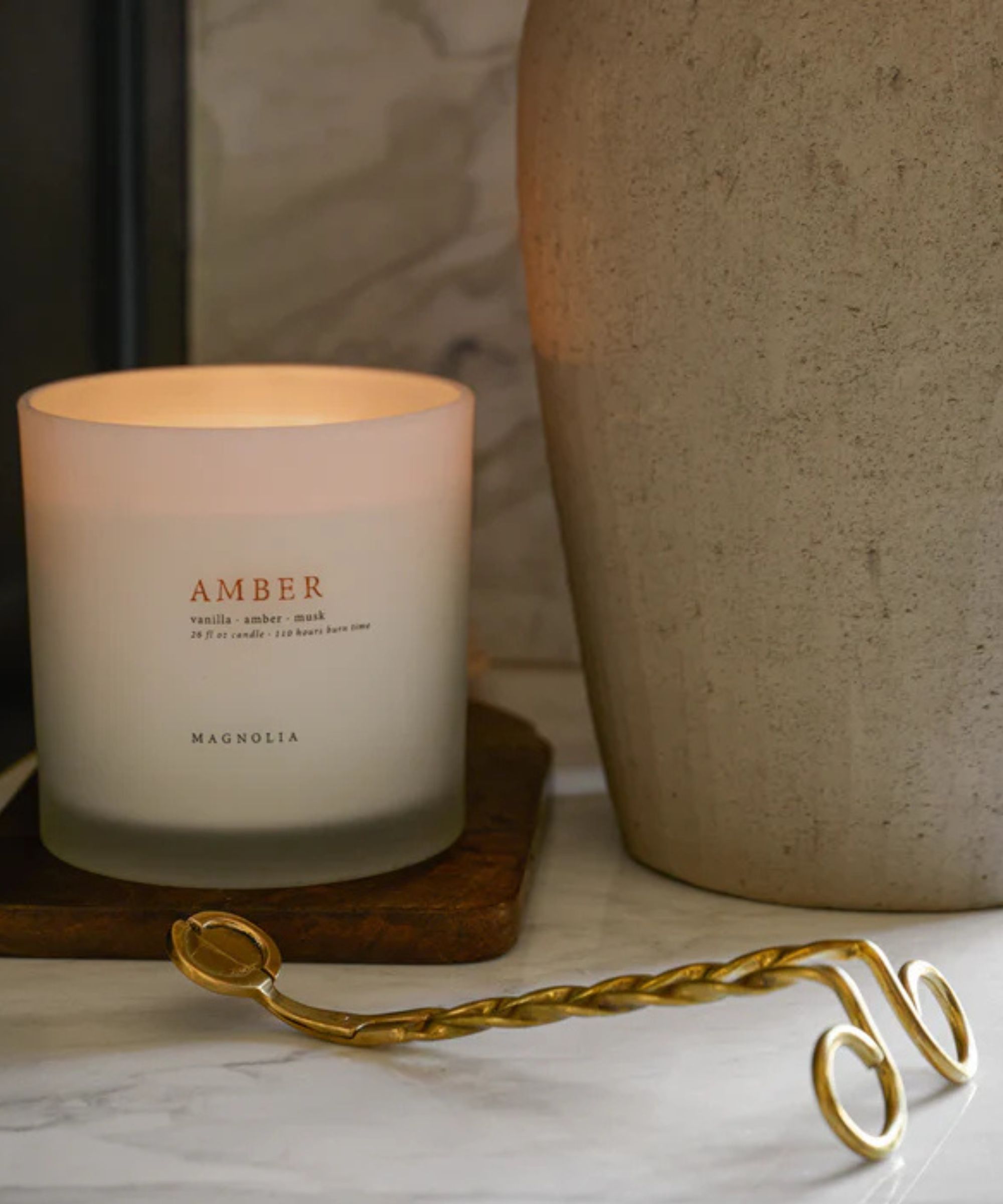 White 'Amber' candle on a wooden coaster with a gold candle snuffer in the foreground