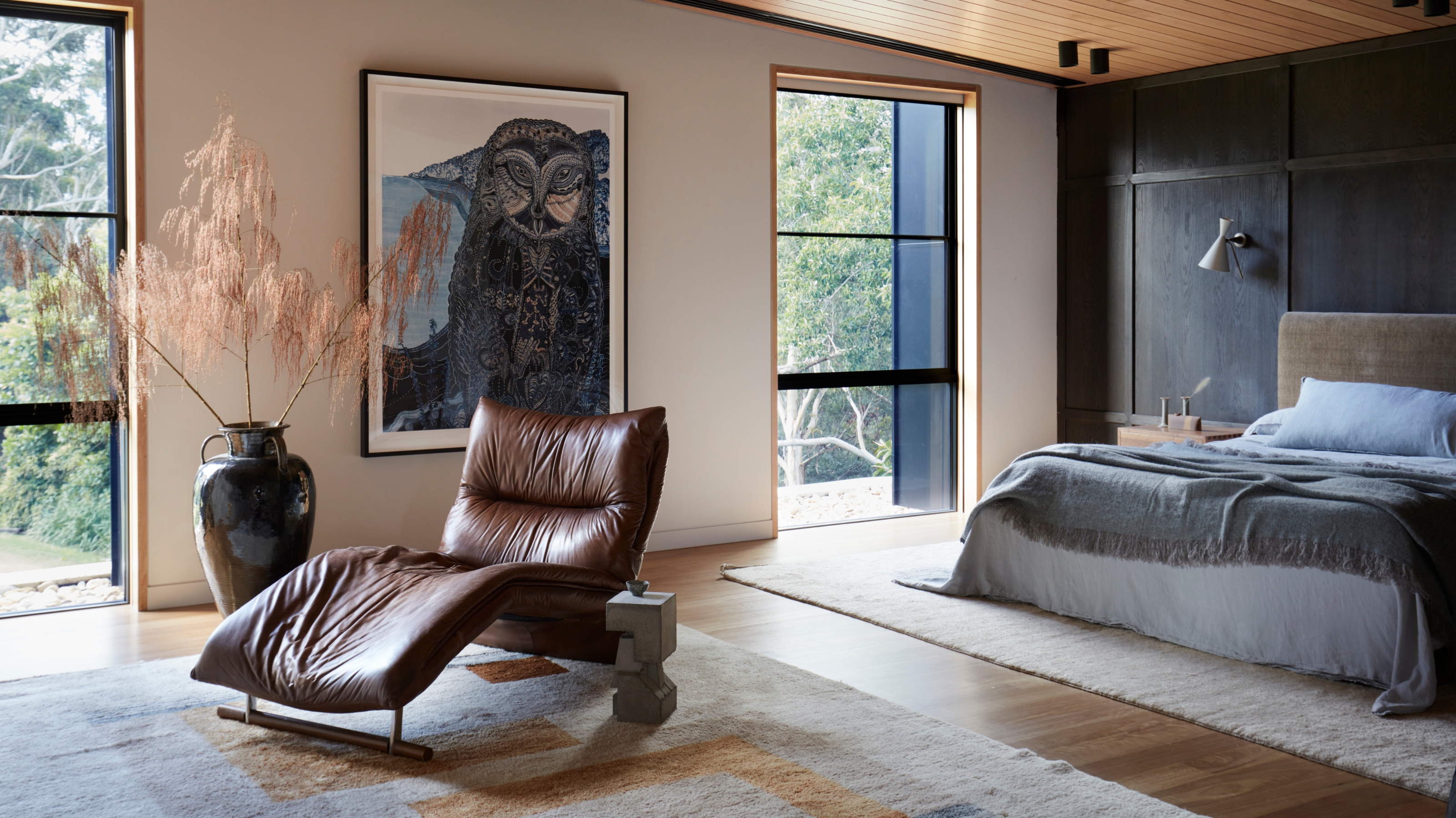 Image of a large bedroom with a dark accent wall and blue-gray bedding. There is a rug under the bed and another rug further out in the sitting area. There is a brown leather lounge chair in the sitting area as well.