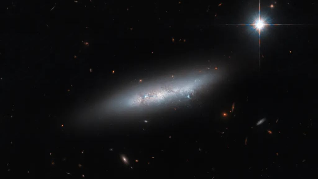 a white shining galaxy hangs in a black space, neighbored by dimmer, smaller, colorful other galaxies the size of shimmering stars. a bright white star gleams in the upper right. An irregular galaxy, NGC 2814, located 85 million light-years from Earth belongs to a group of four &#039;late-type galaxies&#039; collectively known as Holmberg 124. 