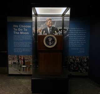 a wooden lectern in a display case stands inside a museum, with a photo of president john f. kennedy in the background.