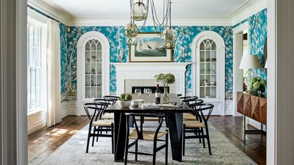 A dining room with turquoise blue and white patterned wallpaper, pendant light with drooping cables, and white arched built-in glass cabinets