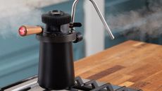 Image of a black stovetop milk steamer on a hob on a wooden kitchen counter. There is steam coming out of the steamer.