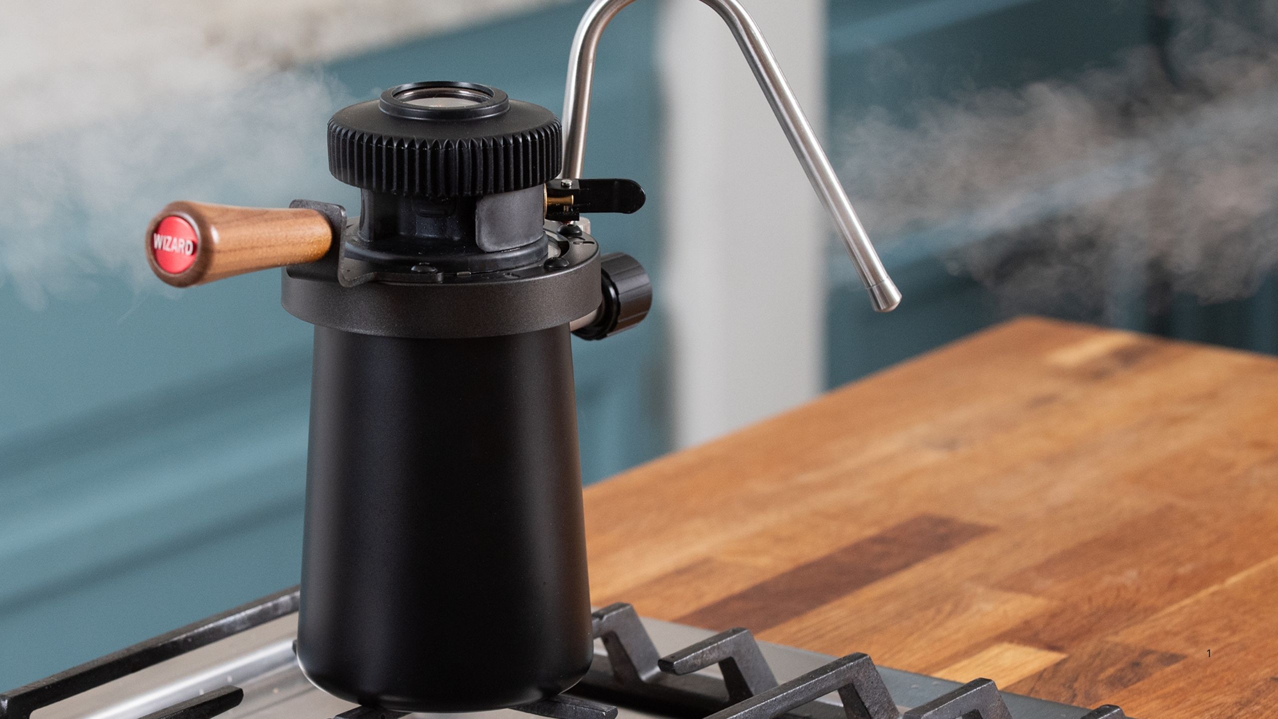Image of a black stovetop milk steamer on a hob on a wooden kitchen counter. There is steam coming out of the steamer.