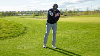 PGA pro John Howells with a golf club across his chest to help him prepare to hit a chip shot from a downhill lie