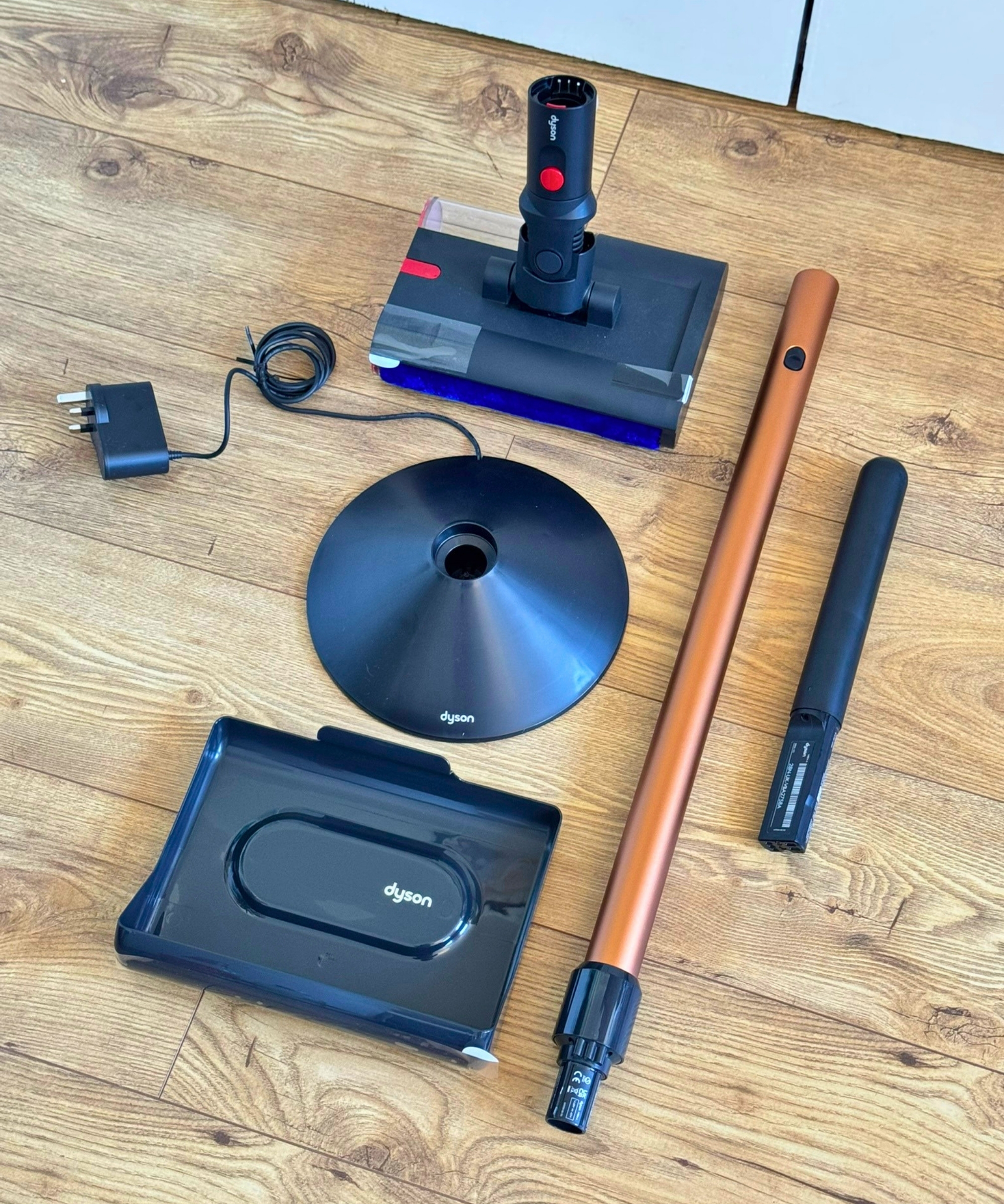 All parts of the Dyson PencilWash on wooden floor, including: floorhead, charging stand, drip tray, wand, handle.