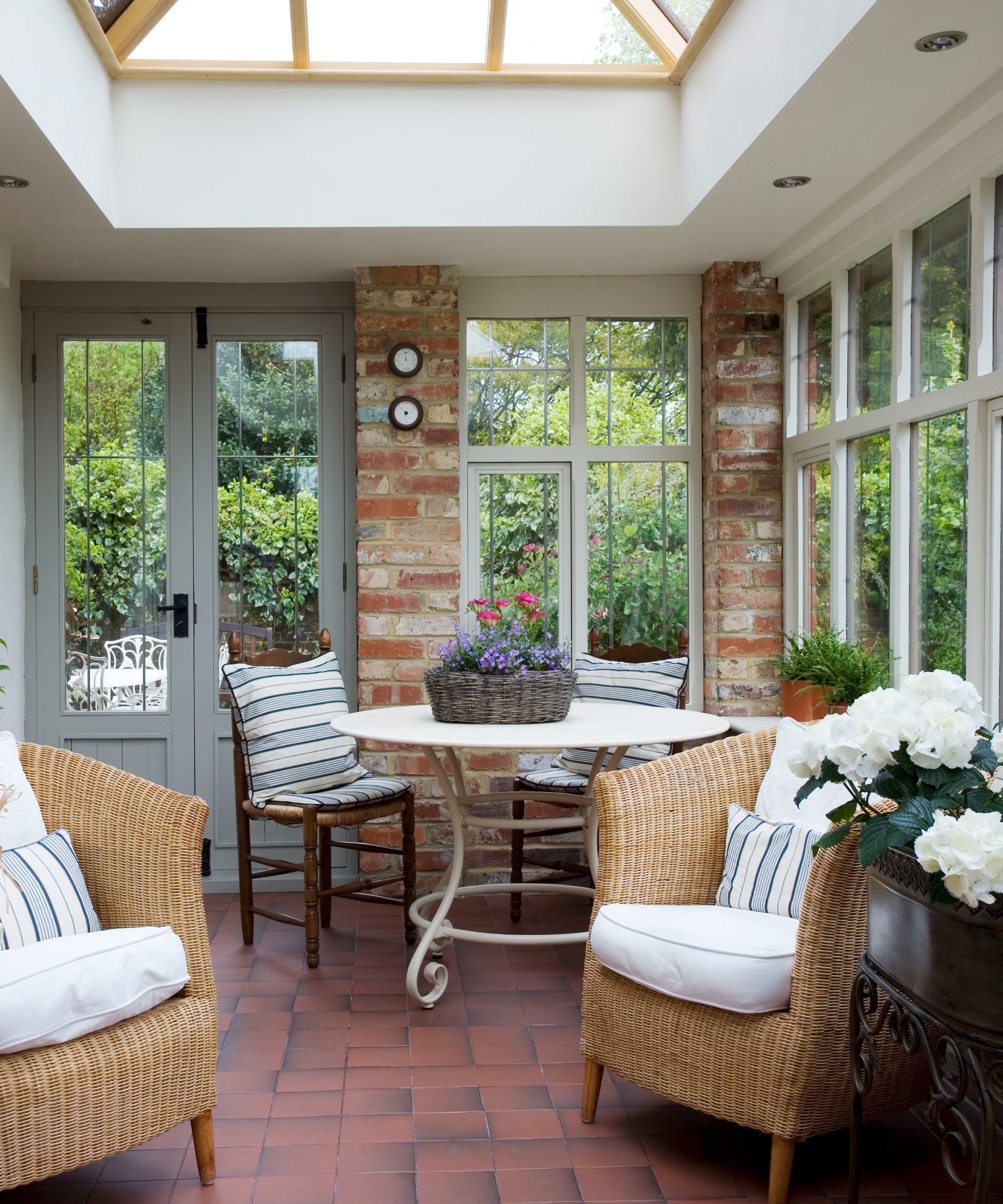 A conservatory with a dark red tiled floor, two rattan armchairs and a round white dining table with two wooden dining chairs