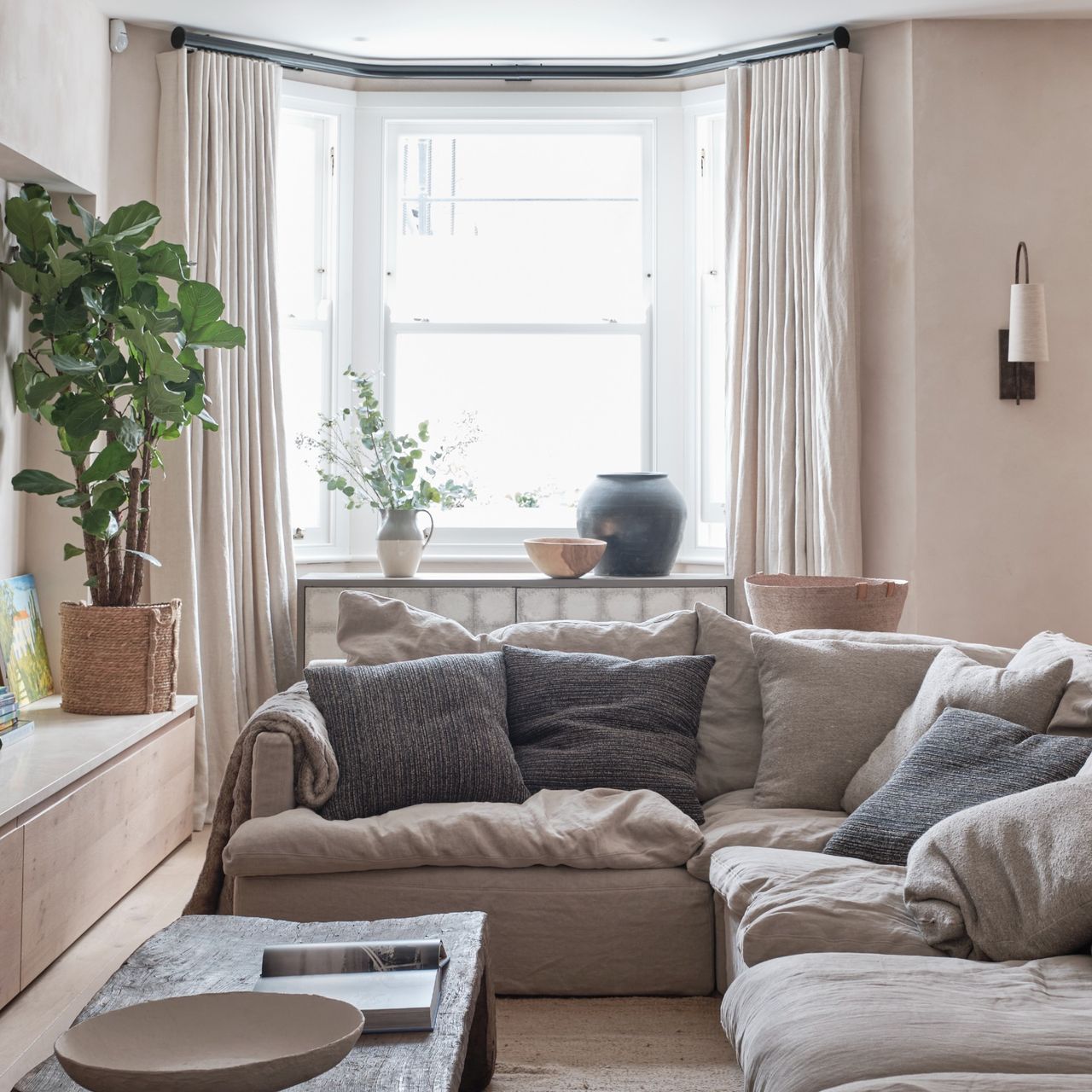 A neutral living room with a corner sofa and full-length curtains framing the bay window