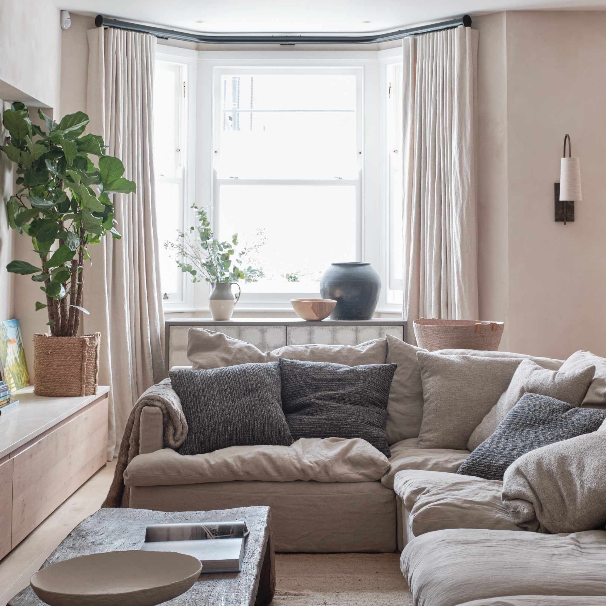 A neutral living room with a corner sofa and full-length curtains framing the bay window
