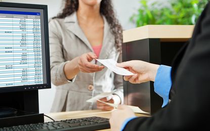 Over-the-shoulder shot of a transaction between a bank teller and a customer in a retail bank. The teller is wearing a black suit and receiving a check from the tan-suited customer over the b