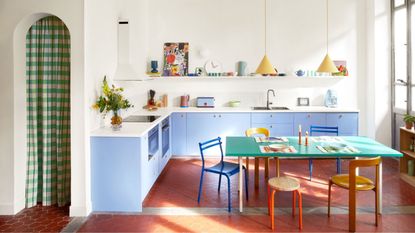 colorful kitchen with arched doorway with checked curtain, light blue cabinetry, white counters, open shelving with colorful decor, red floors, green dining table with colorful chairs, and timber console styled with books