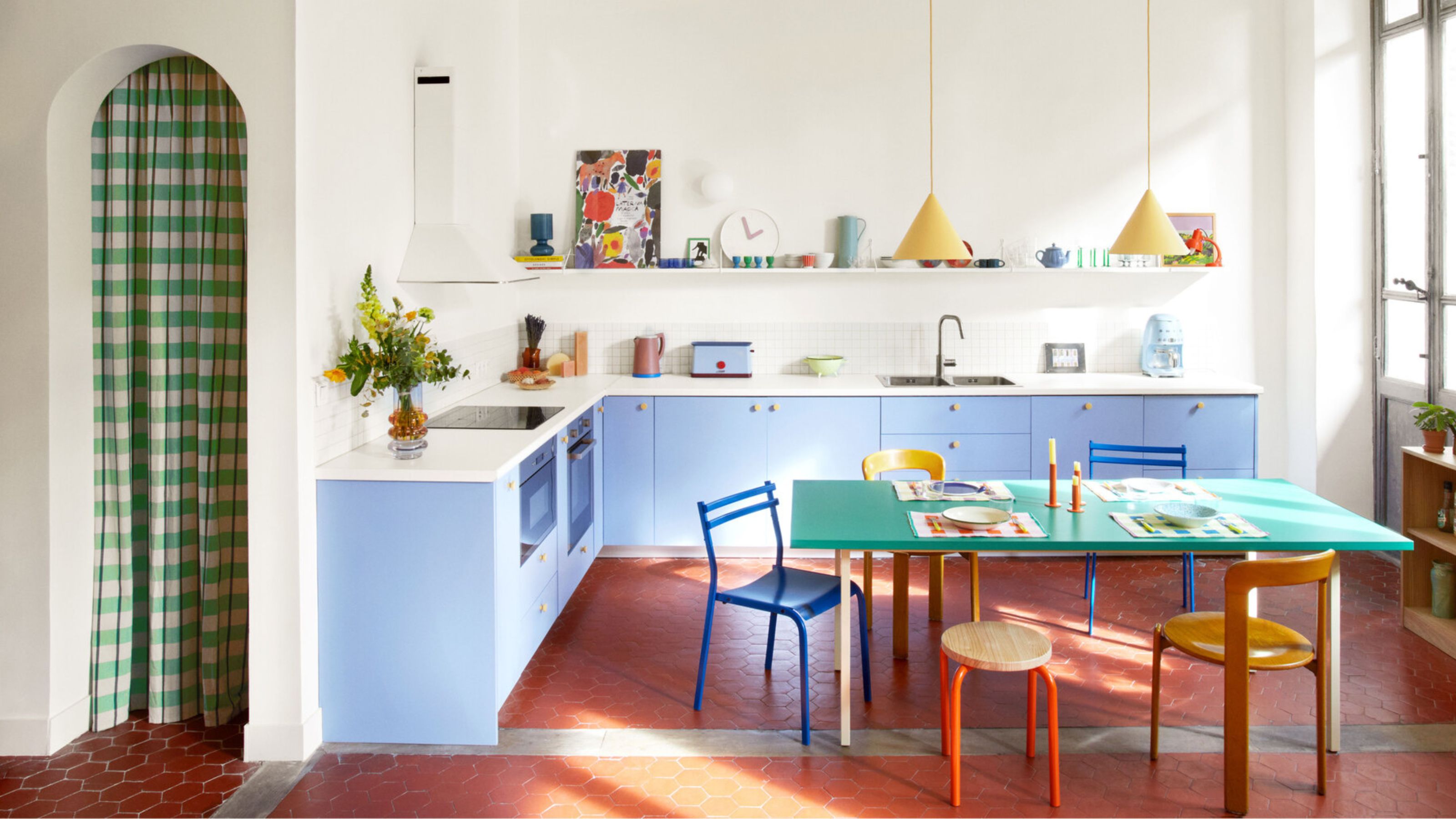 colorful kitchen with arched doorway with checked curtain, light blue cabinetry, white counters, open shelving with colorful decor, red floors, green dining table with colorful chairs, and timber console styled with books