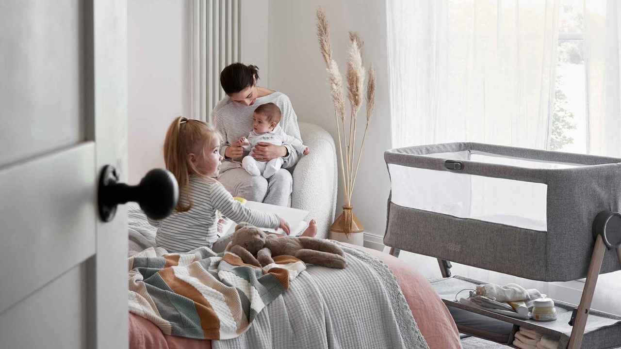 A woman sitting on a white chair with her baby on her lap and a toddler sitting on a bed nearby, with the Lua bedside crib in the background