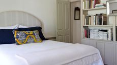 A white bedroom with neutral grey accents. The bed has a grey striped headboard, white bedding and blue and yellow cushions. To the right are some bookshelves and a silver mirror.