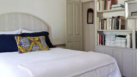 A white bedroom with neutral grey accents. The bed has a grey striped headboard, white bedding and blue and yellow cushions. To the right are some bookshelves and a silver mirror.