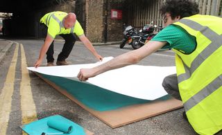 Two men in hi-vis clothing placing a large sheet of paper over a painted piece of wood. They are in the middle of a road