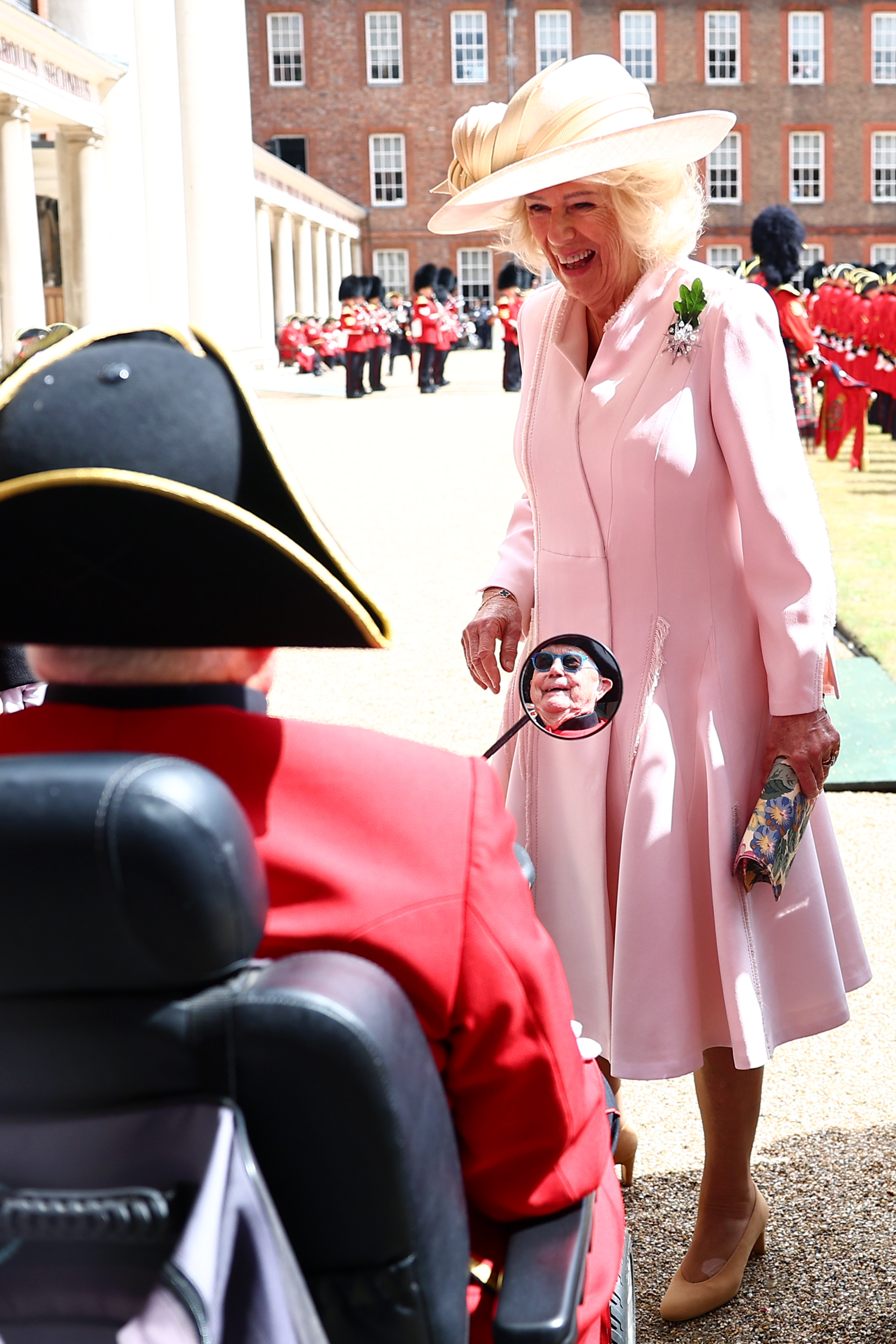 Queen Camilla wearing a pink coat smiling at a man in a scooter