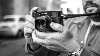 A man holds a vintage camera with both hands, adjusting the focus. The black-and-white image conveys a nostalgic feel, with a blurred car in the background