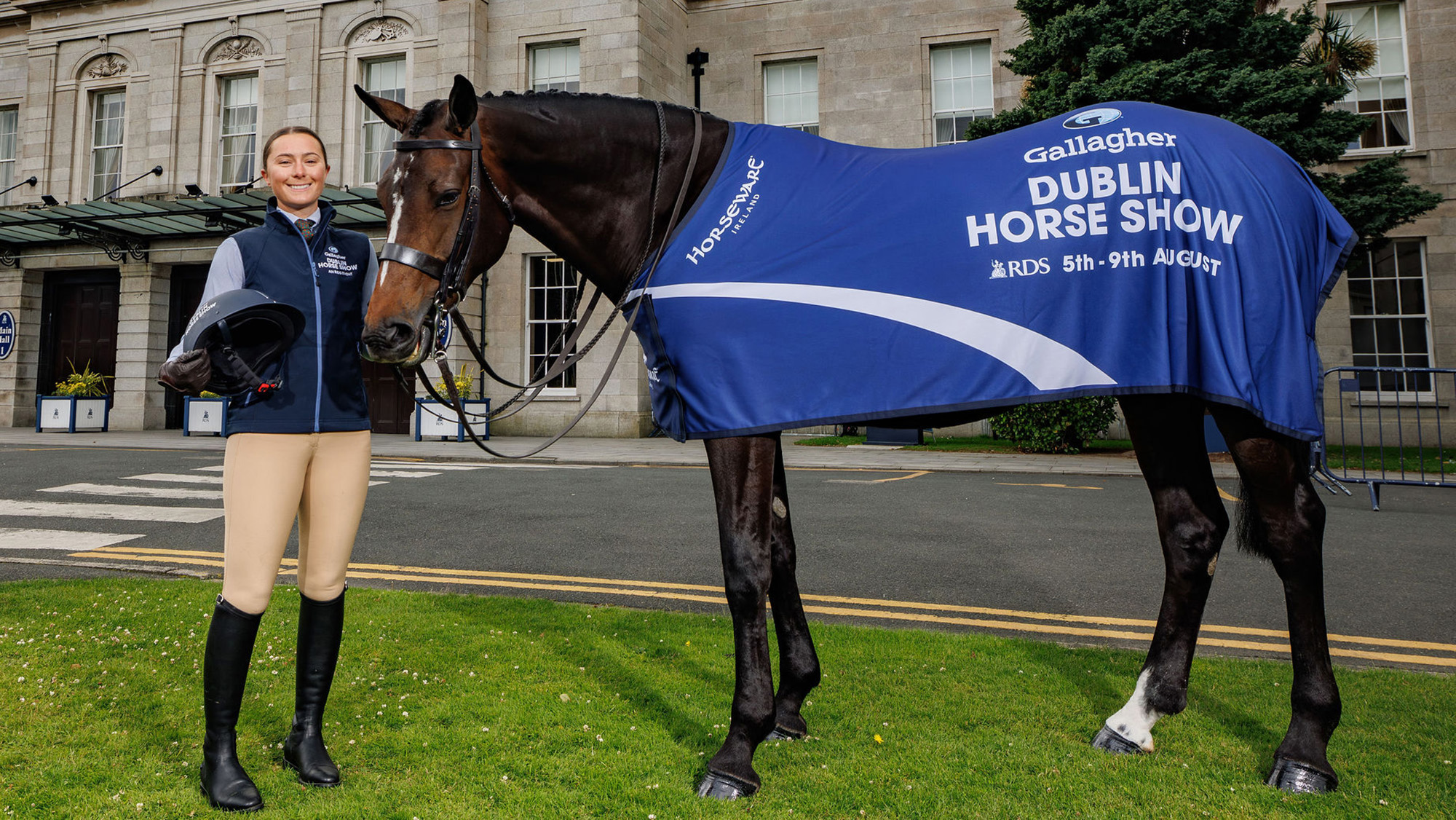 Rachel Moore Rooney stands beside a bay horse with Ruby wearing a blue Dublin Horse Show rug