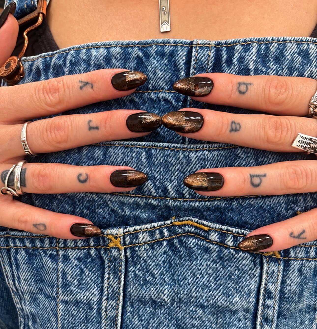 dark brown cat-eye nails with gold shimmer, tiny tattoos and silver rings against denim