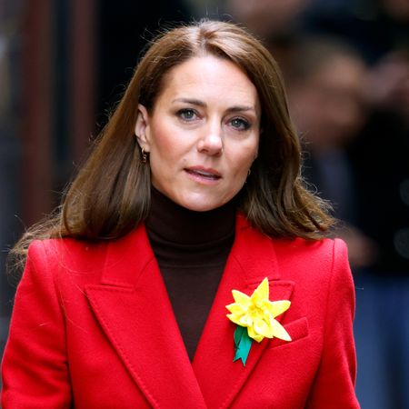 Kate Middleton wears a red coat with a black turtleneck sweater and a yellow daffodil on her lapel, she has long brown hair