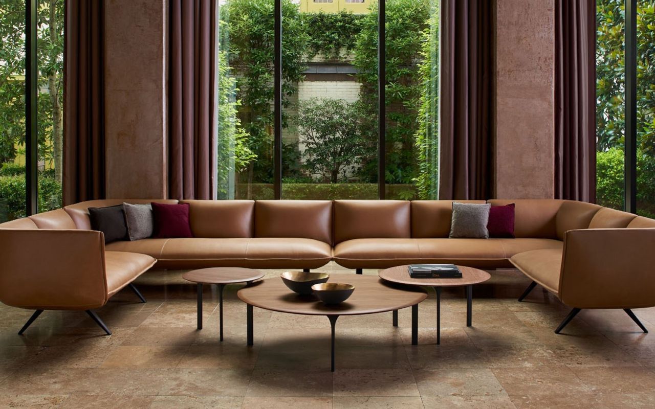 living room with large, minimalist brown sofa, and three wooden coffee tables, in front of large windows overlooking trees