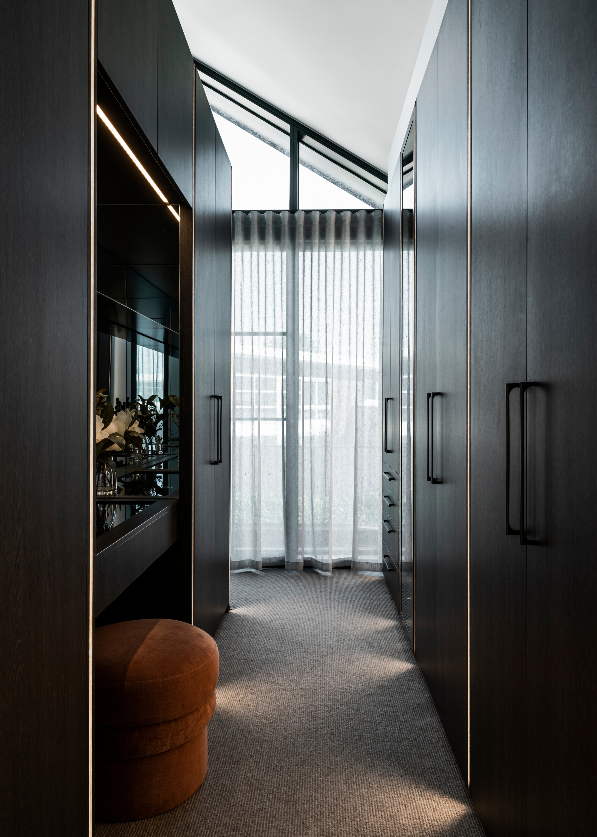A wardrobe with cupboard storage for clothing and a burnt orange pouf by a boudoir
