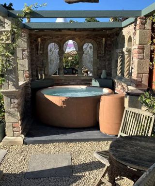 A pergola with a terracotta hot tub, and elegant stone surrounding it, with a small wooden dining set in the corner