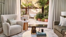 A sunny living room with a boucle chair, green couch, cushions, and a circular coffee table with a vase, candle, and a stack of coffee table books. Behind are large white curtains with an open doorway to a sunny garden behind, with a tiled patio, plants in terracotta pots, and tall palm trees.