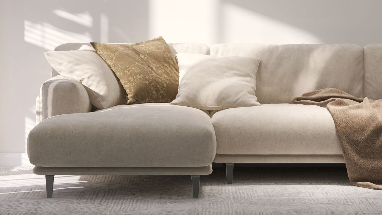 A cream suede sofa with brown cushions and a throw against a white wall